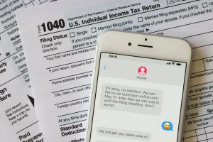 IRS forms, including Form 1040 with a cell phone laying on top of them. The screen of the cell phone shows a text message conversation with Ala Zaki of Elite Accounting, Consulting & Tax.